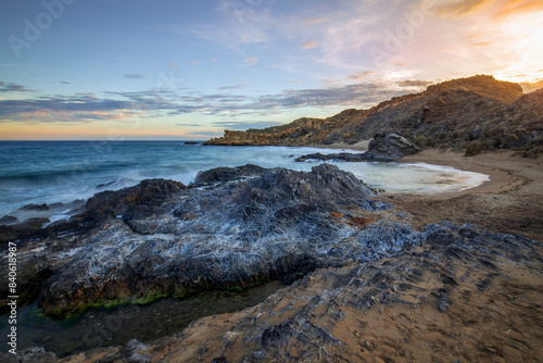 Sunset view of Minas Cove beach in Cabo Cope and Puntas de Calnegre Regional Park, with crystal clear water and rocks in the foreground