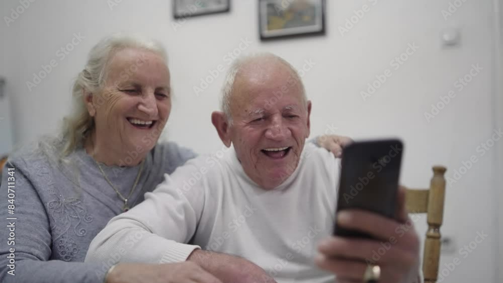 A happy elderly couple enjoys a cheerful video chat on a smartphone at home, laughing and smiling warmly together in a candid moment. A happy elderly couple enjoys a cheerful video chat on a smartphone at home, laughing and smiling warmly together in a candid moment.