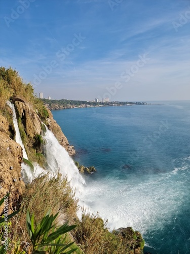 Waterfall in Antalya, Turkiye, water goes down with emerald color into the blue sea under the blue sky, coastline behind it