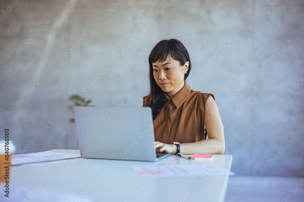 An Asian businesswoman engages with a laptop, wearing professional attire, in a well-lit office environment, reflecting productivity and focus on her professional tasks.