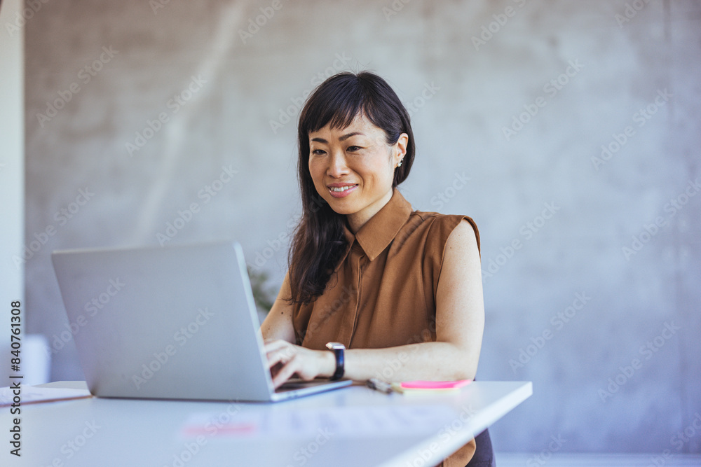 A professional Asian businesswoman engages contentedly with her laptop, donning smart casual attire in a well-lit office setting, embodying efficiency and positivity in the workplace. A professional Asian businesswoman engages contentedly with her laptop, donning smart casual attire in a well-lit office setting, embodying efficiency and positivity in the workplace.