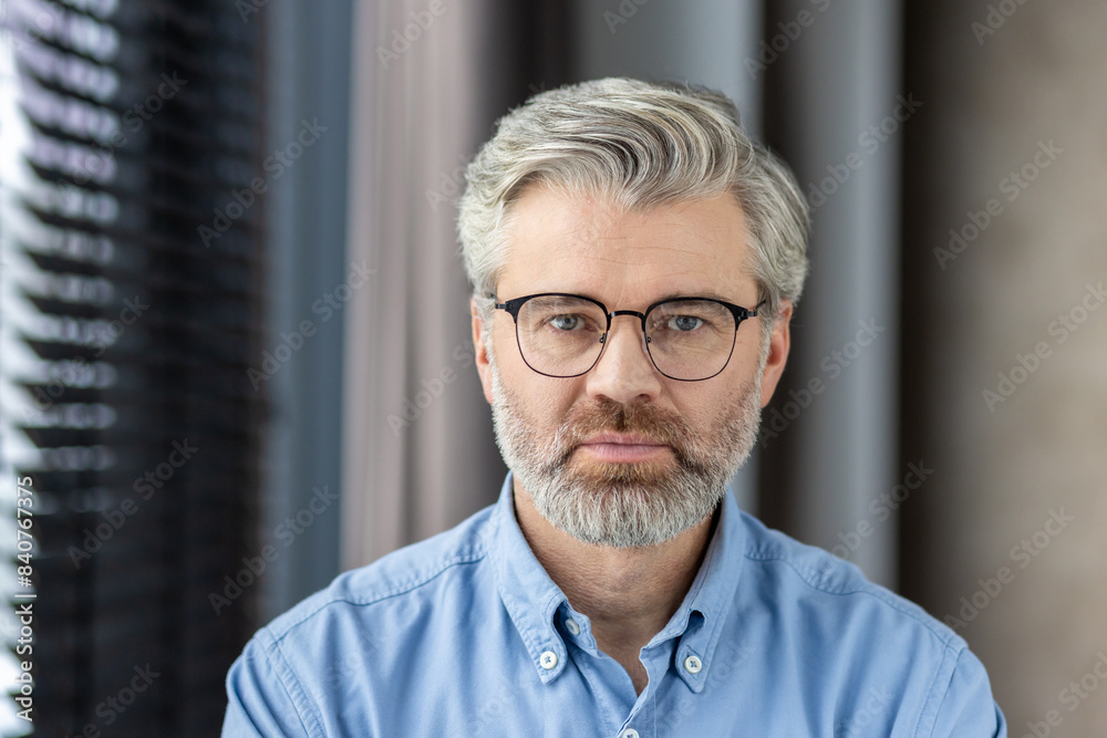 Close-up portrait of a mature man with glasses and beard wearing a blue shirt. Indoors with a blurred background. Close-up portrait of a mature man with glasses and beard wearing a blue shirt. Indoors with a blurred background.