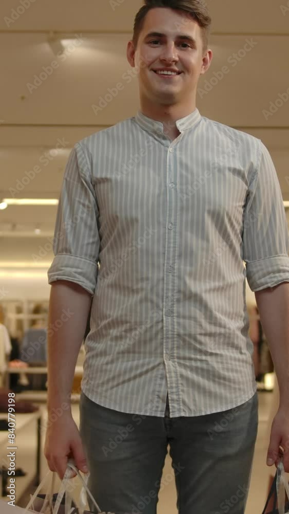 A man with shopping packages approaches the camera with a smile. A young successful guy in a shirt bought in a new modern clothing store. Middle shot. Vertical shot. A man with shopping packages approaches the camera with a smile. A young successful guy in a shirt bought in a new modern clothing store. Middle shot. Vertical shot.