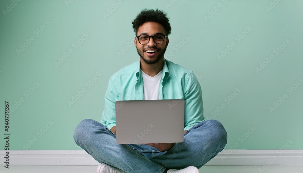 Smiling young man sitting on the floor with her legs crossed holding a laptop computer, pastel background. Student. Male office worker 