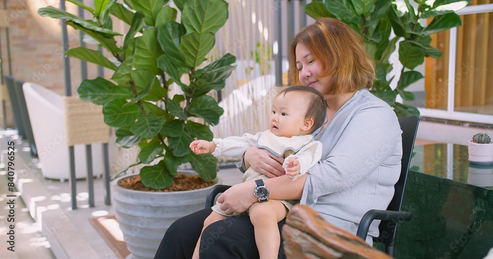Fototapeta premium A loving mother lifts her smiling baby for a kiss in cozy indoor with lush green plants. This heartwarming moment captures the essence of maternal affection joy in a beautiful natural environment