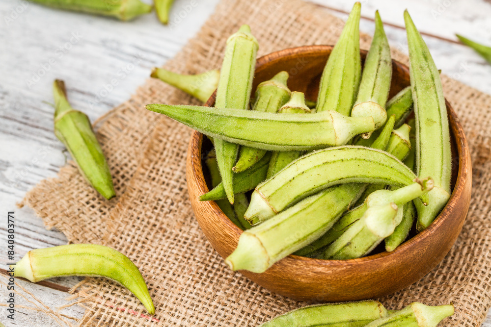 Wooden bowl filled with fresh okra pods placed on a piece of burlap fabric on a rustic wooden white surface. Wooden bowl filled with fresh okra pods placed on a piece of burlap fabric on a rustic wooden white surface.