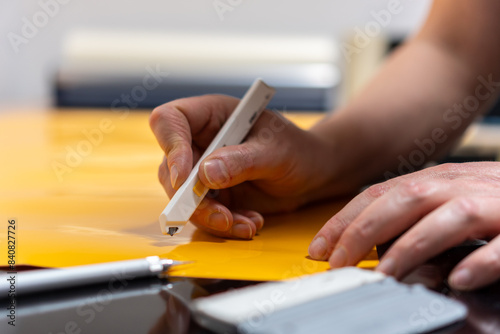 Close-up of an employee's hand holding a utility knife and cutting vinyl film on the table.