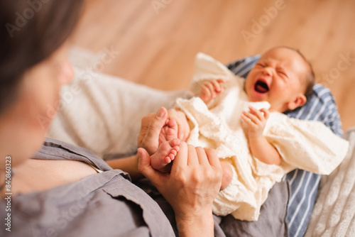 Mother holding crying baby girl holding her feet and toes in room close up. Motherhood. Top view. photo