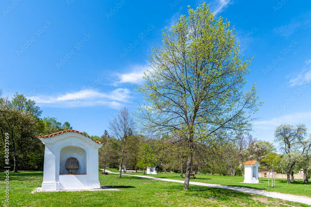 Fototapeta premium Small chapels at Skalka Calvary stand amidst vibrant green trees under a clear blue sky, signaling the arrival of spring. Mnisek pod Brdy, Czechia