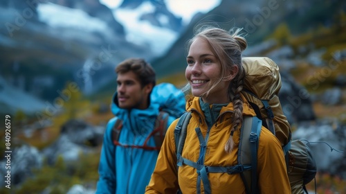 A woman with a backpack is hiking in a mountainous area, smiling, with a male companion slightly out of focus in the background
