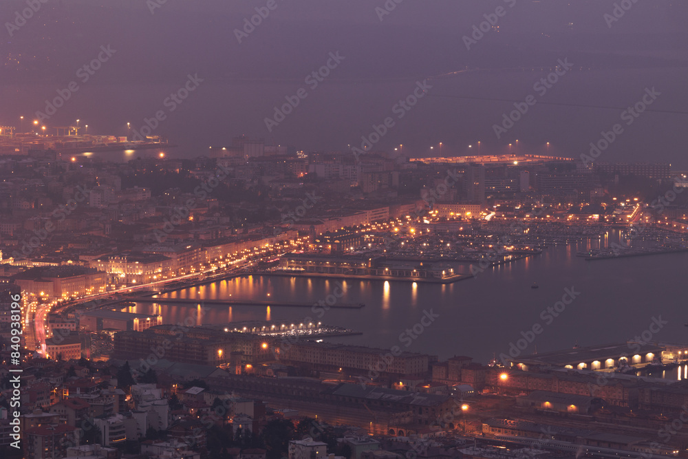 Fototapeta premium vista in primo piano di parte della città di Trieste nella zona centrale e del porto che si affaccia sul mare Adriatico e il golfo, visto dall'alto, di sera, con le luci delle strade accese