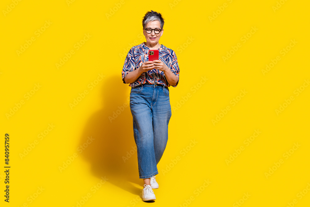 Full length photo of lovely senior lady walk hold device dressed stylish colorful garment isolated on yellow color background Full length photo of lovely senior lady walk hold device dressed stylish colorful garment isolated on yellow color background