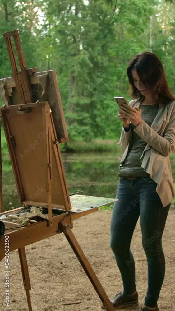 A girl artist, next to an easel and a palette, communicates with her friends on a social network in a smartphone. In full growth. Park and lake in the background. Vertical shot. A girl artist, next to an easel and a palette, communicates with her friends on a social network in a smartphone. In full growth. Park and lake in the background. Vertical shot.