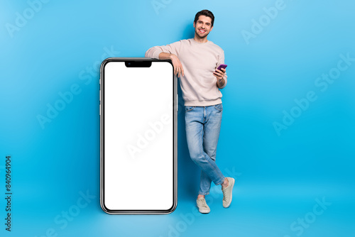 Full size photo of nice young man use smart phone empty space placard wear beige sweatshirt isolated on blue color background