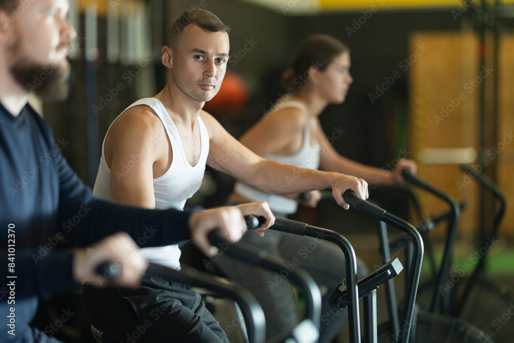 Fototapeta premium Concentrated athletic young adult man intensely working out on stationary air bicycle in fitness gym. Healthy active lifestyle concept