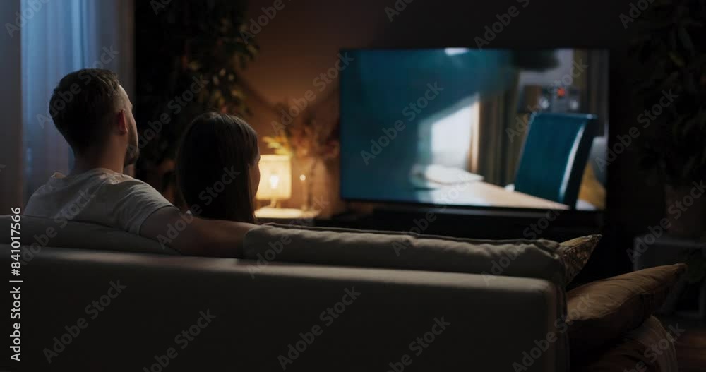 From behind young woman and man sit snuggled up together on couch watching romantic movie occasionally steal glanceing at each other hold hands hugging feeling content and relaxed.
