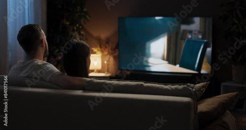 From behind young woman and man sit snuggled up together on couch watching romantic movie occasionally steal glanceing at each other hold hands hugging feeling content and relaxed.