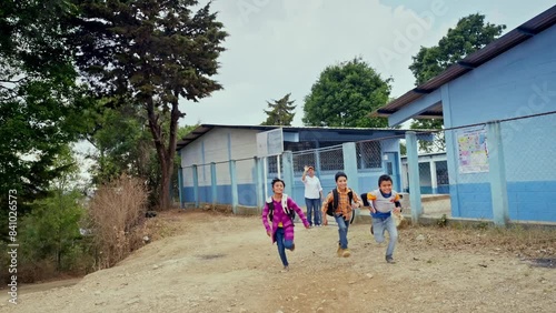 A group of happy children run in front of the school and the teacher says goodbye to them with a cordial greeting.