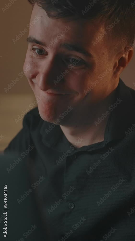 Caucasian man smiles cutely while talking in an online conference. Confident attractive man writhes a cute face, wriggles in the interior of a fashionable cozy apartment, close-up. Vertical shot. Caucasian man smiles cutely while talking in an online conference. Confident attractive man writhes a cute face, wriggles in the interior of a fashionable cozy apartment, close-up. Vertical shot.