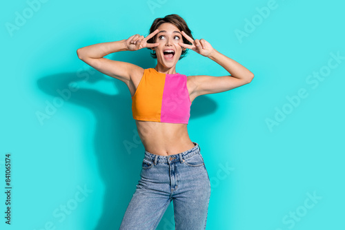 Photo of flirty excited woman wear pink orange top showing two v-signs looking empty space isolated teal color background photo