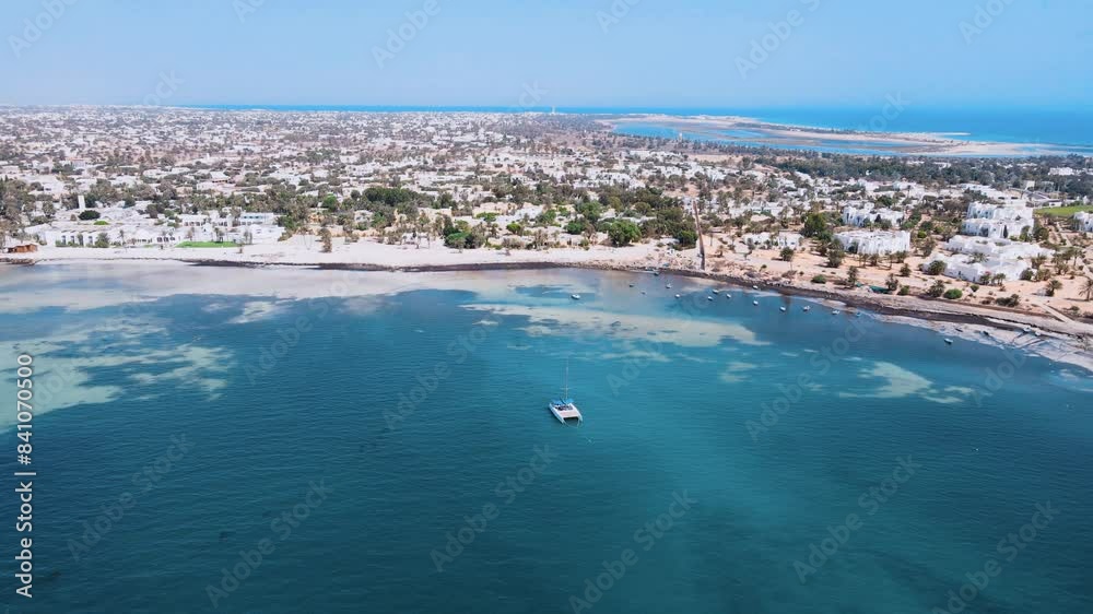 An aerial view of a sailboat is floating on a large body of water, surrounded by a beautiful sandy shores. beach with palm trees and houses nearby. at Tunisia Djerba An aerial view of a sailboat is floating on a large body of water, surrounded by a beautiful sandy shores. beach with palm trees and houses nearby. at Tunisia Djerba