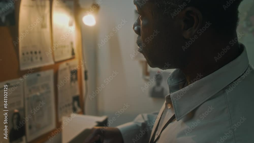 Medium close-up tilt shot of African American investigator working in office, pouring cup of coffee from glass jug and drinking, while staring at pin board with crime photos Medium close-up tilt shot of African American investigator working in office, pouring cup of coffee from glass jug and drinking, while staring at pin board with crime photos