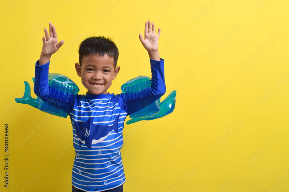Portrait of a cute little Asian boy wearing a swimsuit posing with both hands raised isolated on a yellow background. Summer vacation concept. With Copy space Portrait of a cute little Asian boy wearing a swimsuit posing with both hands raised isolated on a yellow background. Summer vacation concept. With Copy space