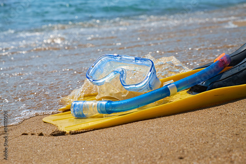 A pair of bright yellow flippers and a blue snorkel lie on the soft sand of a tropical beach. The turquoise ocean waves gently lap at the shore in the background.