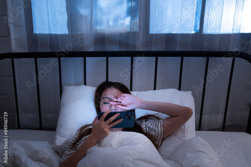 Asian young woman lying in bed at night holding a smartphone and covering her eye from the screen brightness of blue light unhealthy lifestyle before sleep