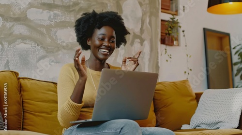 Gleeful, attractive Black lady utilizing laptop, engaging in virtual meeting, communicating with hand gestures while seated on cozy couch in her abode. Concept of communication.