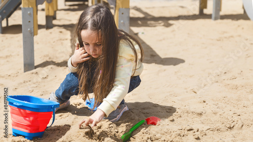 Pretty child girl playing in sand on outdoor playground. Beautiful baby in jeans trousers having fun on sunny warm summer day. Child with colorful sand toys. Healthy active baby outdoors plays games © Mariia