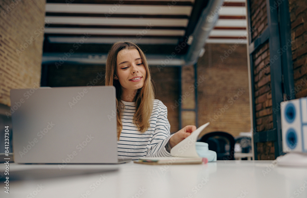 Young female freelancer working on laptop while sitting in modern coworking and look in notepad Young female freelancer working on laptop while sitting in modern coworking and look in notepad