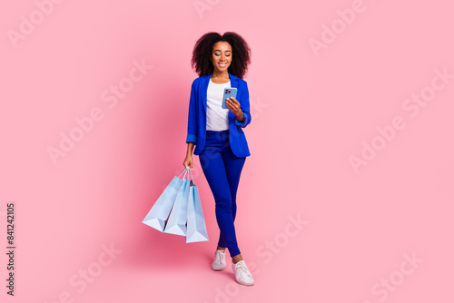 Full length photo of friendly cheerful woman wear blue jacket holding shopping bags look at smartphone isolated on pink color background