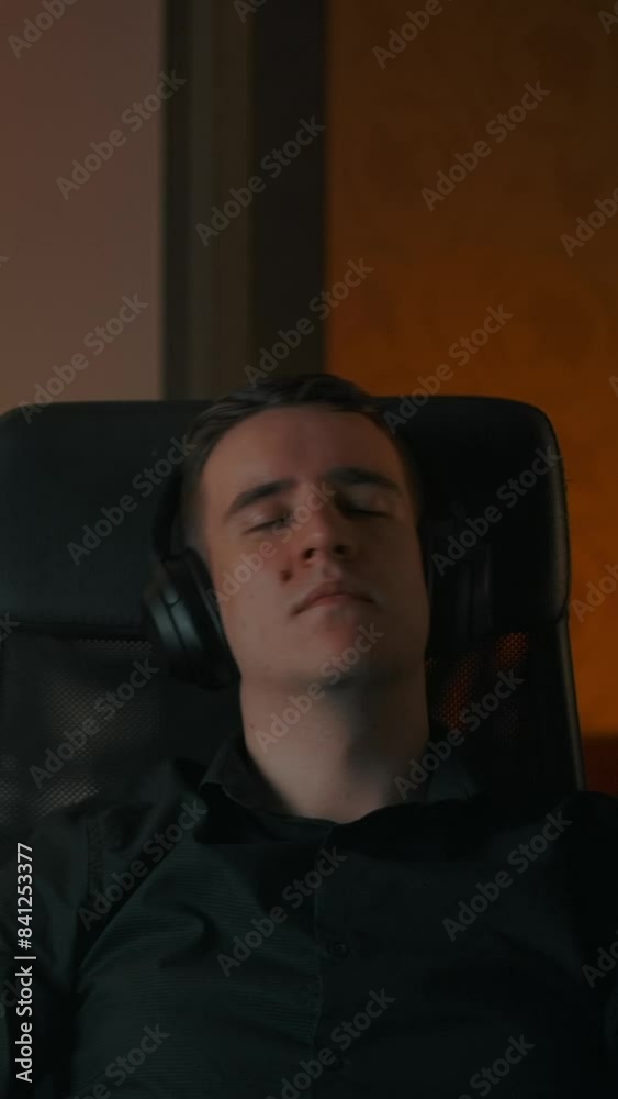 Caucasian man relaxing on armchair in cozy home while listening music. Big earphones, melodic music for meditation and relaxing behind laptop. Night look Vertical shot.