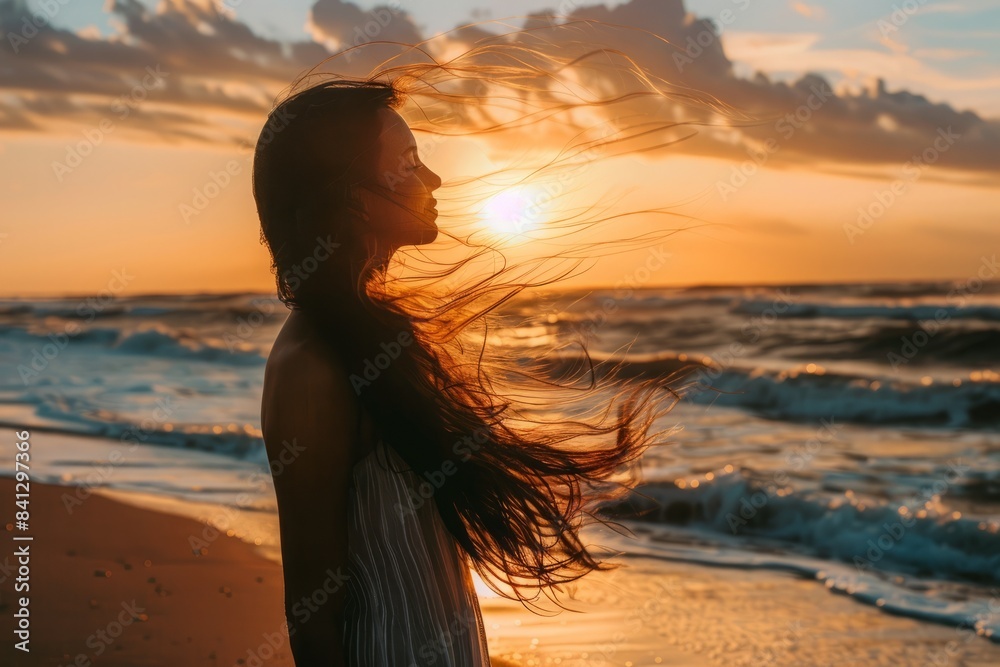 A beautiful woman with long hair blowing in the sea breeze, standing on the beach at sunset A beautiful woman with long hair blowing in the sea breeze, standing on the beach at sunset