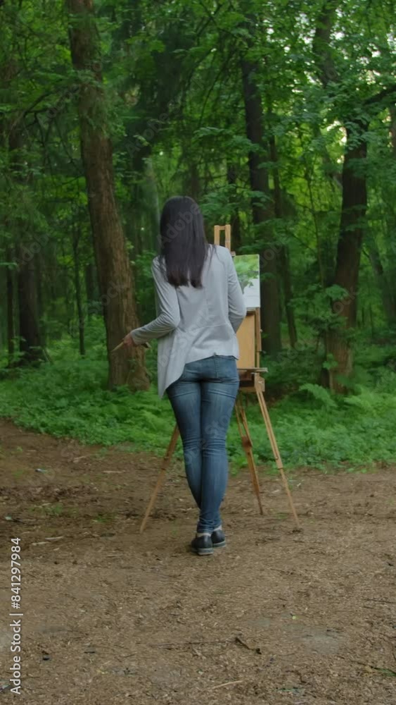 Girl draws a landscape, standing in front of an easel in a park. General plan beautiful women in jeans and a jacket on the background of the lake and tree trunks. Wide shot. Vertical shot. Girl draws a landscape, standing in front of an easel in a park. General plan beautiful women in jeans and a jacket on the background of the lake and tree trunks. Wide shot. Vertical shot.