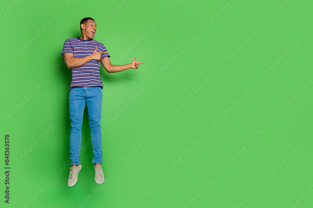 Full body portrait of nice young man direct fingers empty space jump wear t-shirt isolated on green color background Full body portrait of nice young man direct fingers empty space jump wear t-shirt isolated on green color background