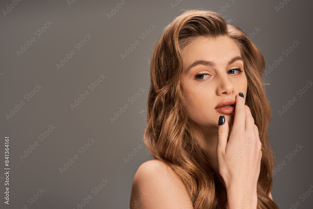 A young woman with long, wavy hair delicately touches her face, exuding beauty and grace. A young woman with long, wavy hair delicately touches her face, exuding beauty and grace.