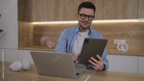 A man, wearing glasses, works from home using a laptop and a tablet. He is a freelancer in the field of online education.
