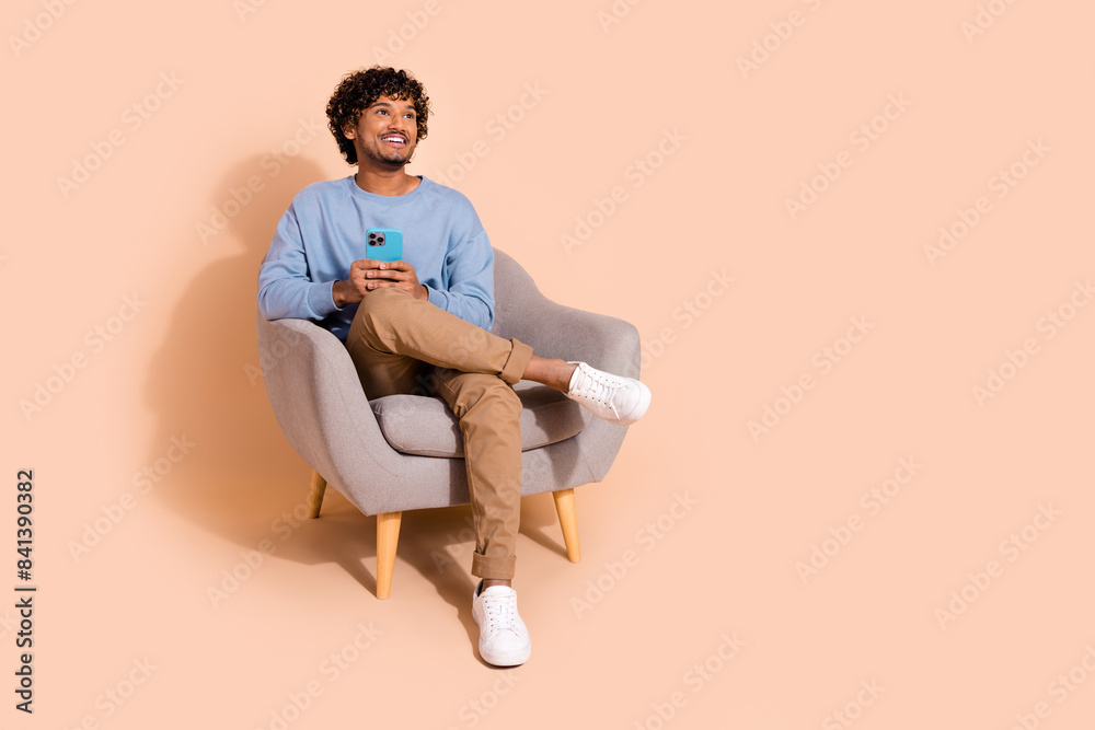 Full length photo of positive dreamy guy dressed blue sweatshirt texting modern device looking empty space isolated beige color background Full length photo of positive dreamy guy dressed blue sweatshirt texting modern device looking empty space isolated beige color background