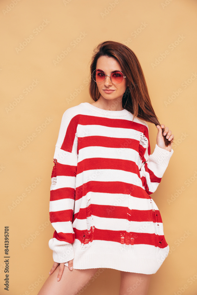 Young beautiful smiling female in trendy striped white and red sweater with holes. Carefree woman isolated on beige wall in studio. Positive model having fun. Cheerful and happy