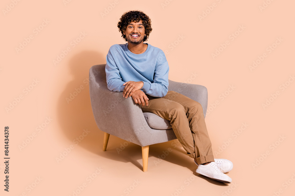 Full length photo of funky cheerful guy dressed blue sweatshirt sitting arm chair smiling isolated beige color background Full length photo of funky cheerful guy dressed blue sweatshirt sitting arm chair smiling isolated beige color background