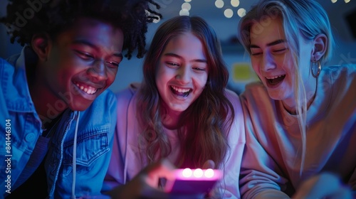 Three diverse teenagers laugh together while looking at a phone screen.  The image is lit with vibrant, colorful lights.