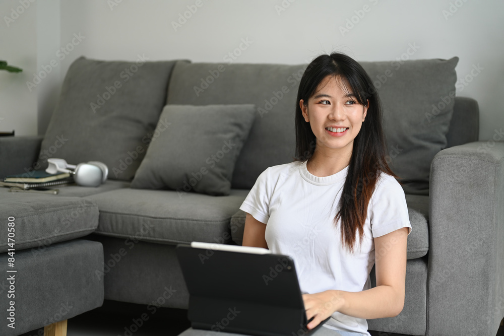 Charming young Asian woman sitting in living room with digital tablet and looking away Charming young Asian woman sitting in living room with digital tablet and looking away