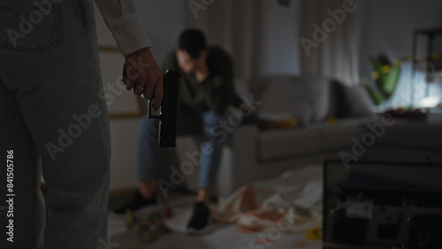 In a dimly lit room, a worried man sits on the couch as a woman stealthily holds a gun, suggesting tension and danger indoors.