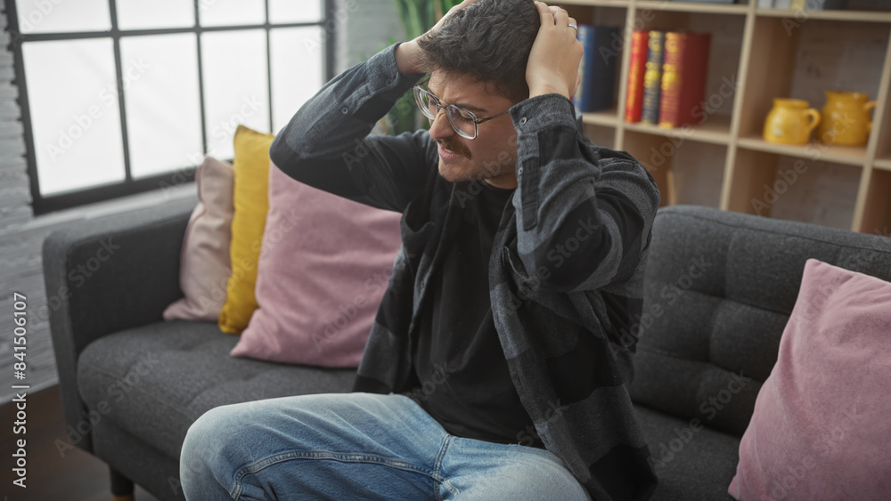 Stressed young man sitting on couch indoors with hands on head in a modern apartment. Stressed young man sitting on couch indoors with hands on head in a modern apartment.