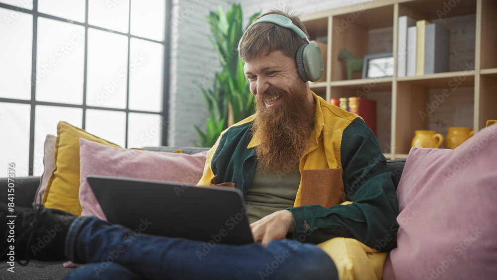 Redheaded bearded man smiling using laptop with headphones indoors, sitting on colorful couch by window. Redheaded bearded man smiling using laptop with headphones indoors, sitting on colorful couch by window.