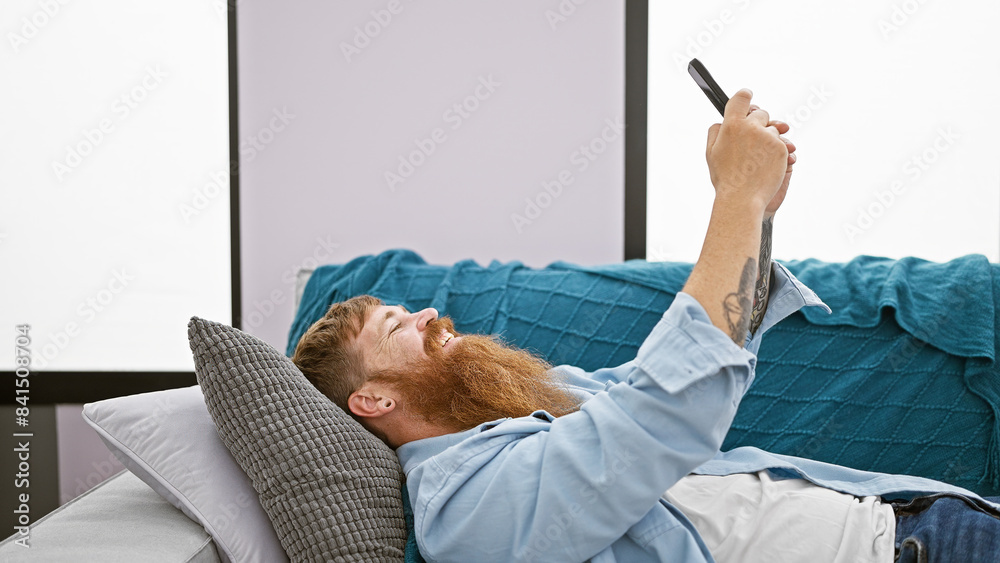 Cheerful young redhead man joyfully typing away messages on his smartphone, lying comfortably on the sofa, lost in the fun world of internet, right in the heart of his cozy living room at home
