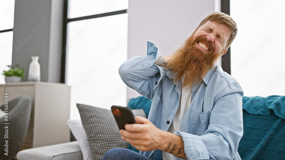 Stressed redhead guy nursing neck pain on sofa while engrossed in phone at home Stressed redhead guy nursing neck pain on sofa while engrossed in phone at home