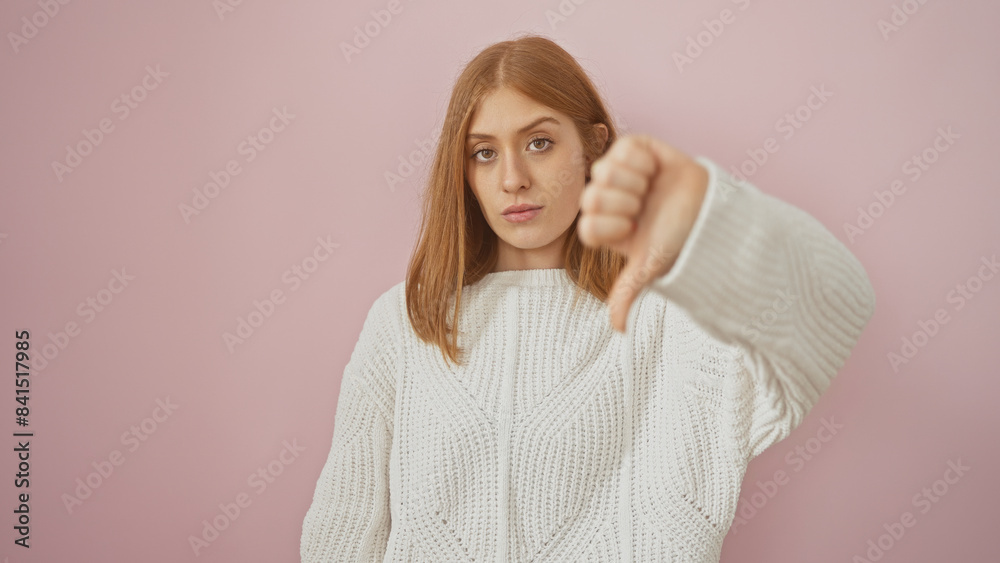 Caucasian redhead woman in white sweater giving thumbs down gesture against pink background. Caucasian redhead woman in white sweater giving thumbs down gesture against pink background.